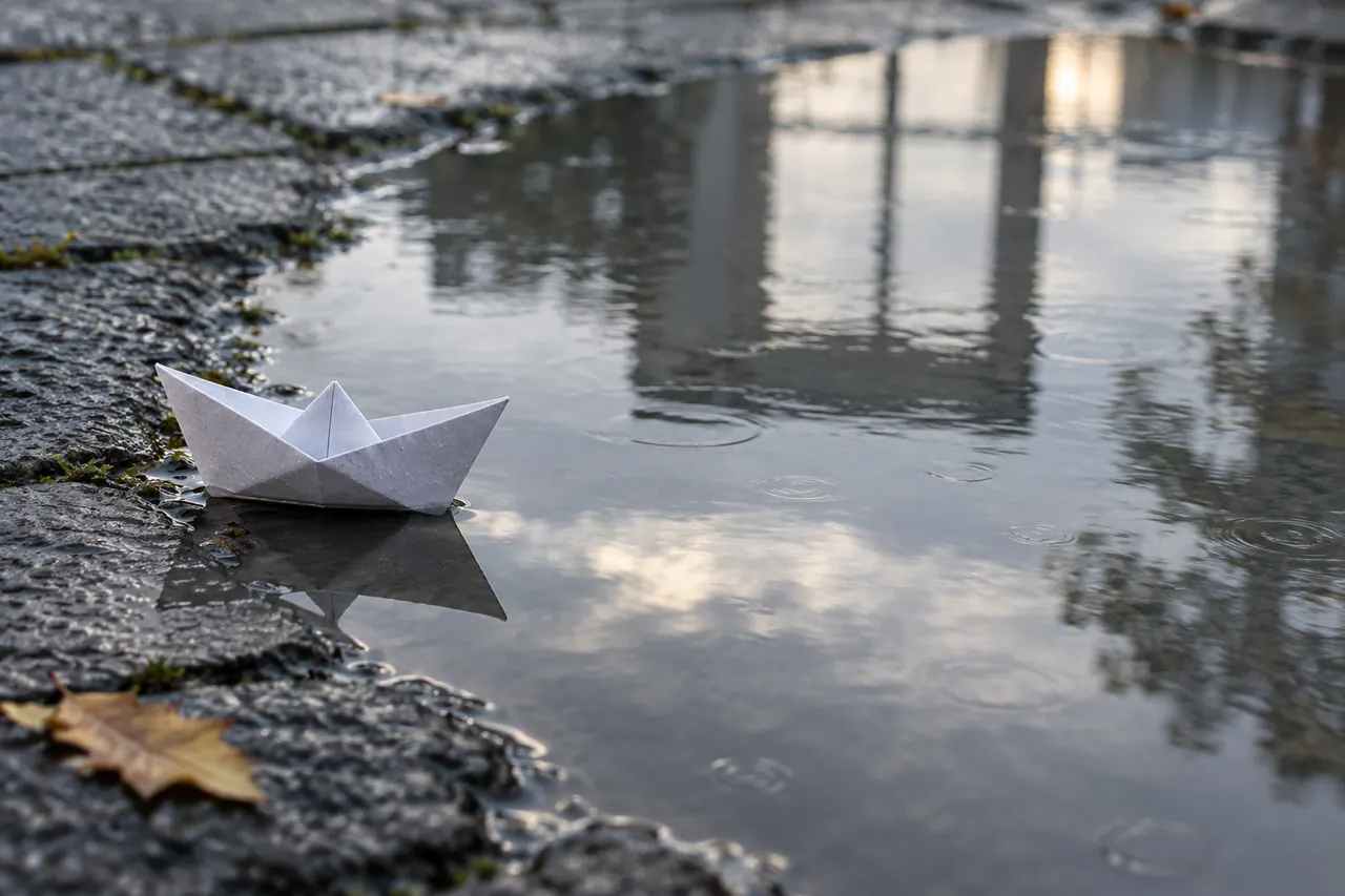 A small white folded paper boat at the left edge of a shallow puddle on dark gray paving after rain.